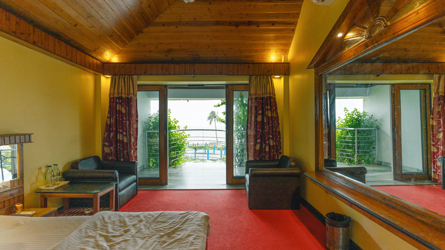 A bedroom with large windows leading to a balcony, 2 armchairs, a table, a mirror, wooden ceiling, and a king size bed at Hotel Seagull Digha.